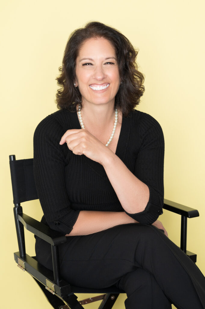 Linda Carmo smiles while she sits in a directors chair in a black outfit in front of a light yellow background.